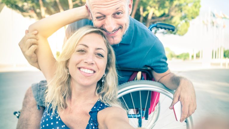 Mujer rubia tomandose un selfie junto a su esposo en silla de ruedas