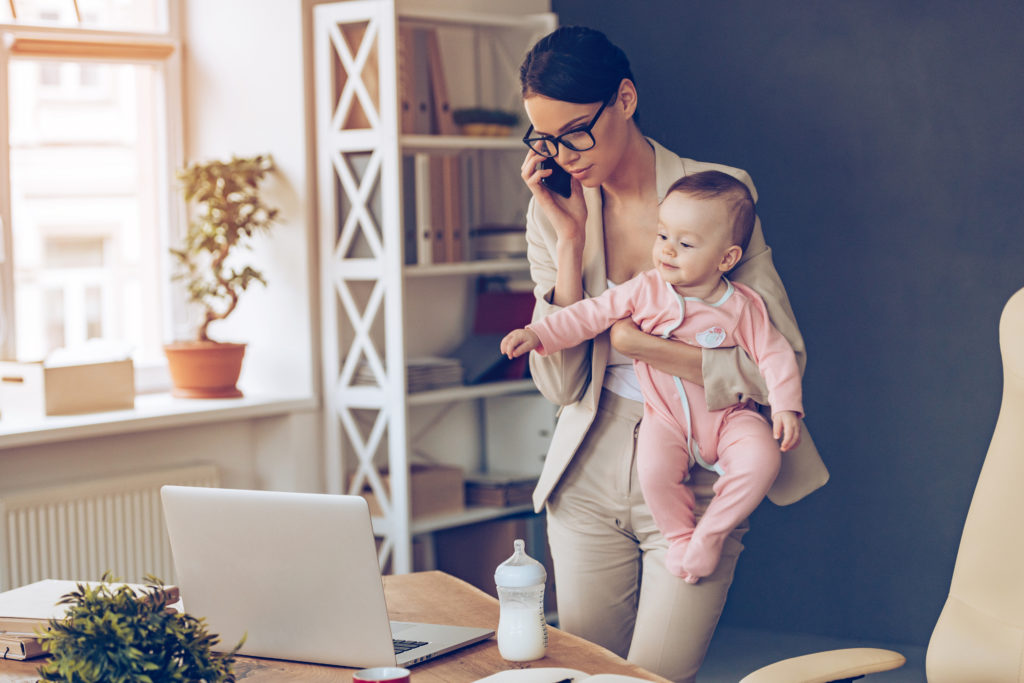 joven empresaria hablando por teléfono móvil y mirando la computadora portátil mientras está de pie con su bebé en su lugar de trabajo