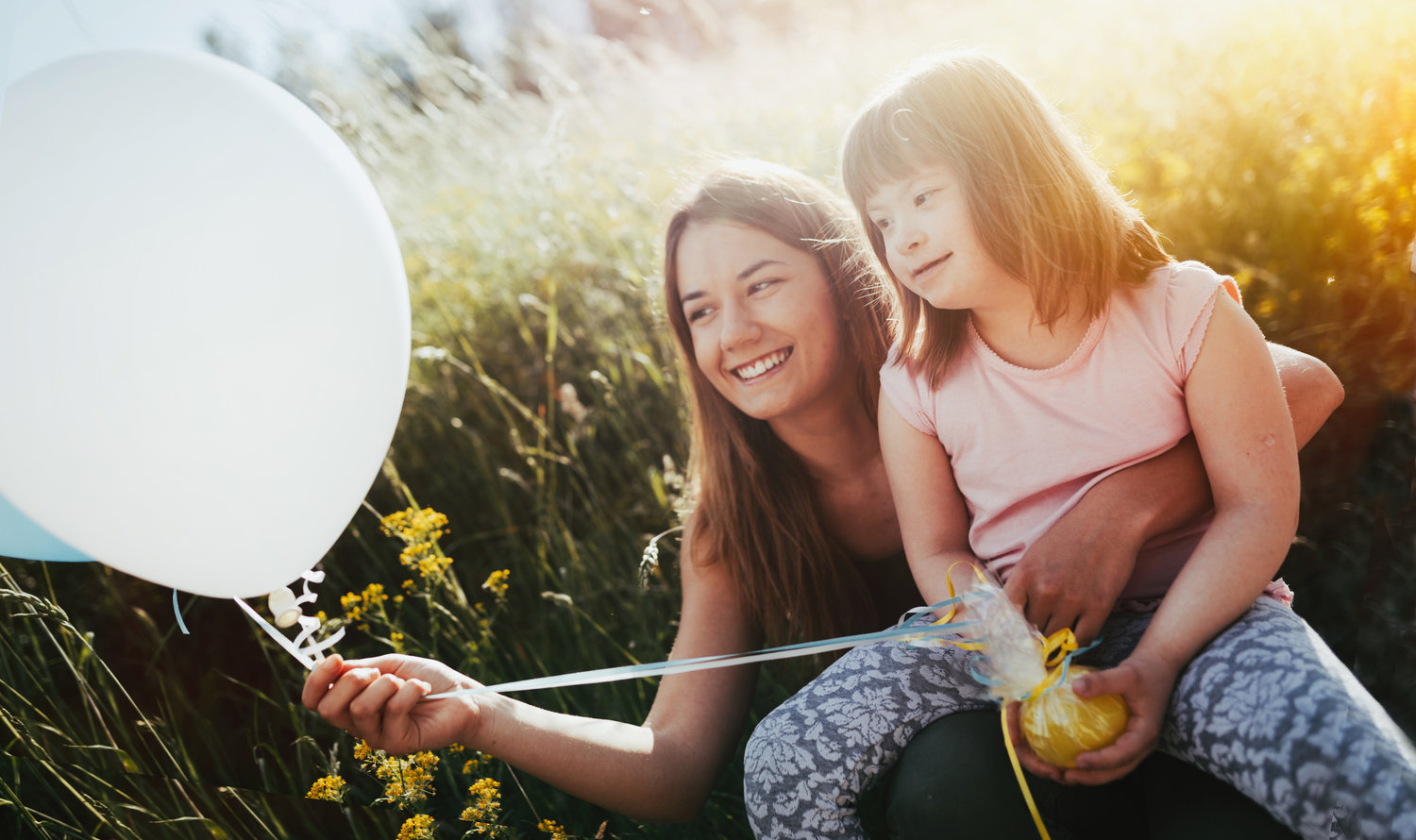 Niña con sindrome de down sentada en los regasos de su madres en un campo que sostiene globos blancos y celestes