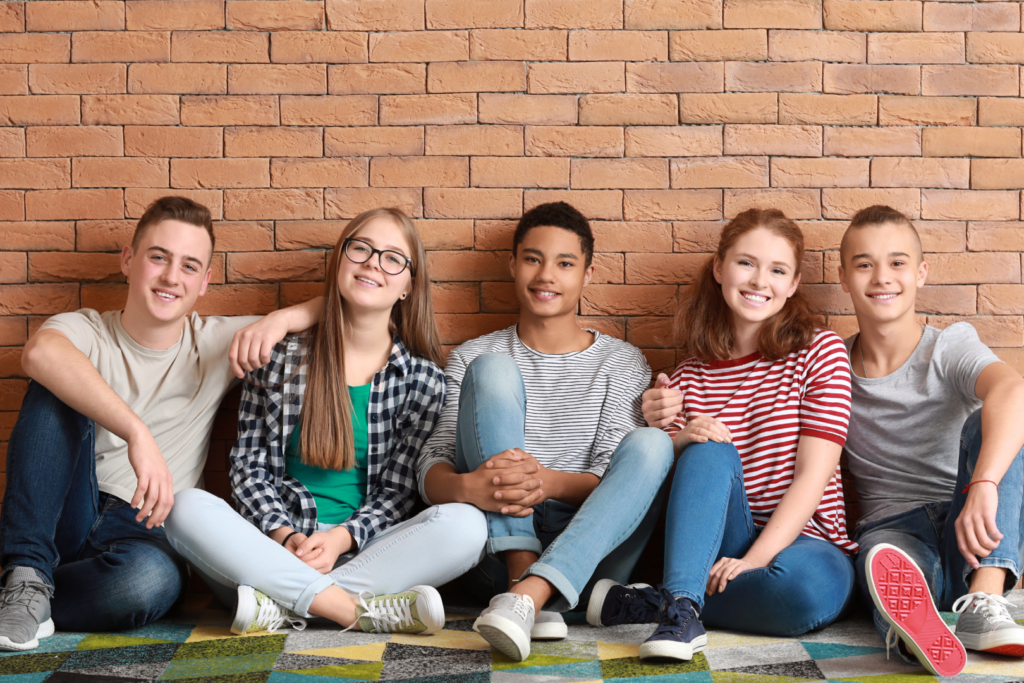 Grupo de 5 jovenes sentados en el piso sonriendo a la camara, detras hay una pared de ladrillos claros