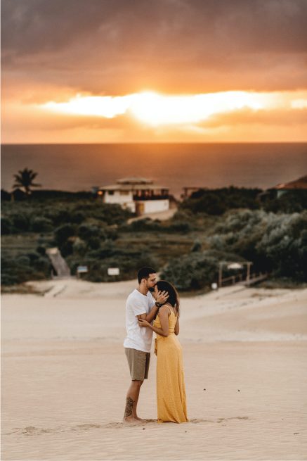 Fotografía de una pareja en la playa con ropa fresca juntos, el hombre le está dando un beso en la frente a su esposa. Se ve una cabañas al fondo y un atardecer