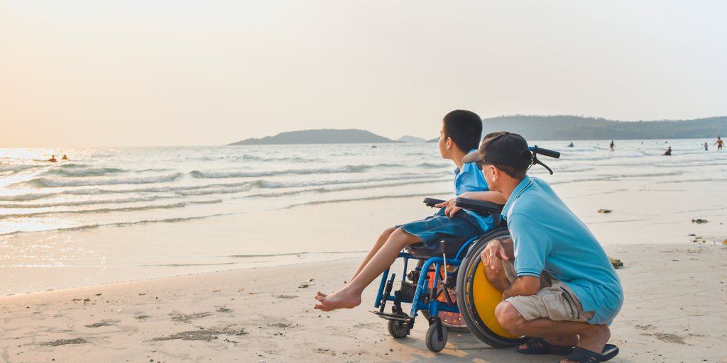 Padre junto a niño en silla de rueda viendo el mar en la playa