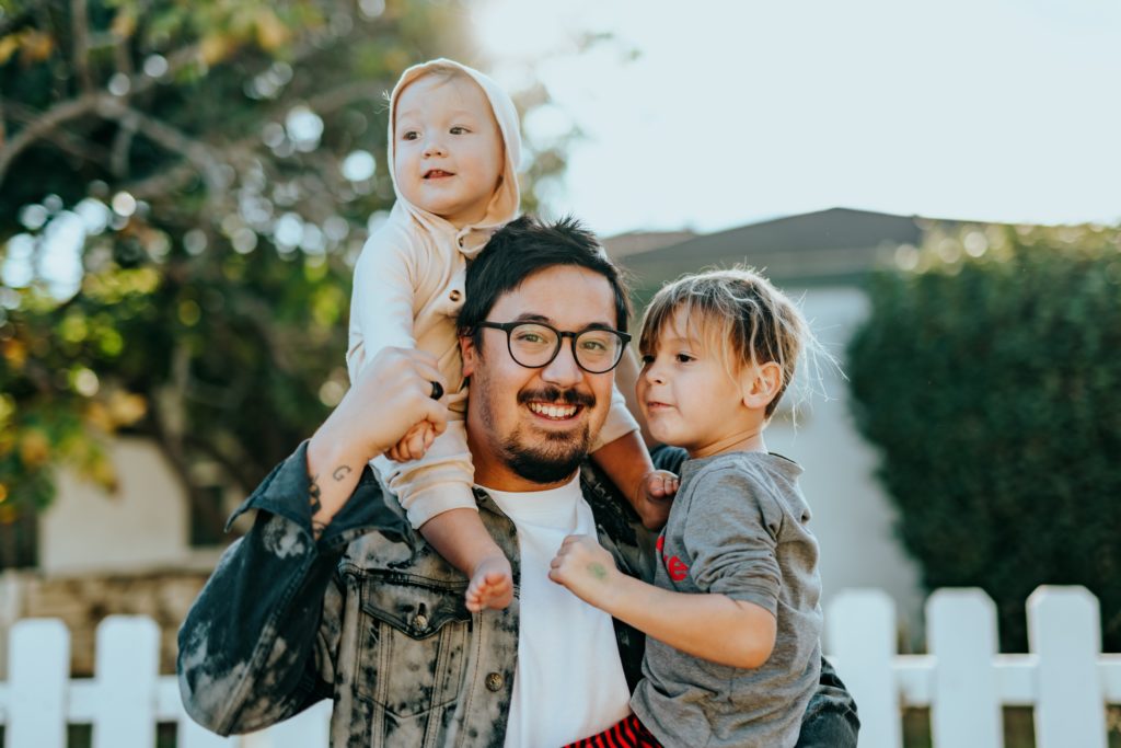 Padre joven con dos hijos en brazos sonriendo en un jardin